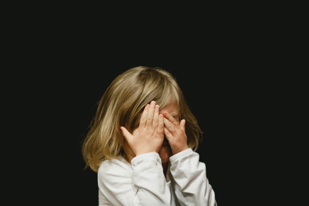 両手で顔を覆って目をふさぐ、白い服を着た小さな子どもの写真